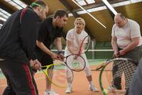 Eine Frau und drei Männer bilden einen Halbkreis in einer Sporthalle. Alle haben eine Tennisschläger in der Hand. Die sehende Frau erläutert den blinden und sehbehinderten Männern den Umgnag mit dem Tennisschläger.