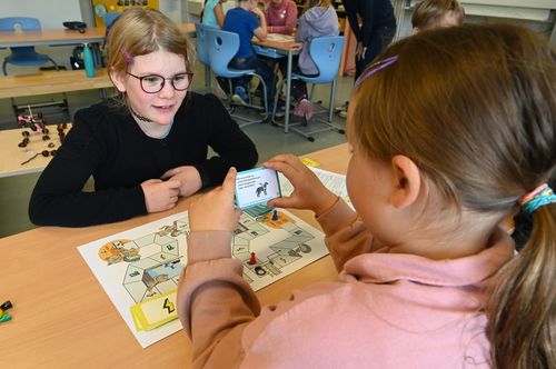 Zwei Mädchen testen das Brettspiel "Rund ums Auge". Sie sitzen in einem Klassenraum mit ihren Mitschülerinnen und Mitschülern. 