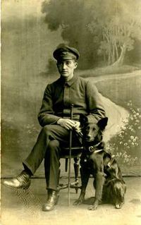 Ein Schwarz/Weiß-Bild. Ein Mann in Uniform sitzt auf einem Stuhl. Neben ihm sitzt sein Blindenführhund.