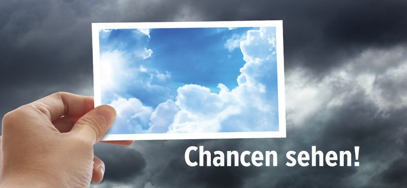 Motiv 2016: Es zeigt einen grauen, wolkigen Himmel. Im Vordergrund hält eine Hand vom linken unteren Bildrand her eine Postkarte in die Bildmitte. Die Karte zeigt einen blauen Himmel mit weißen Wolken. Unter der Postkarte steht das Motto „Chancen sehe