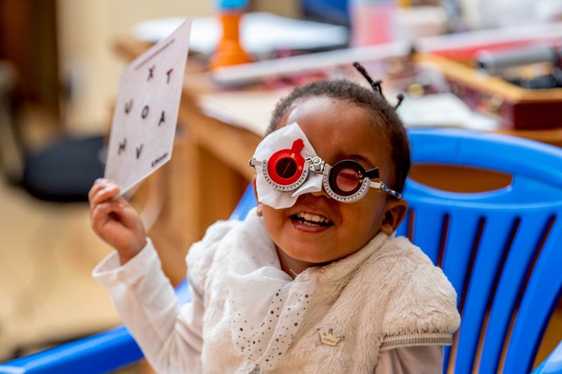 Ein Kleinkind mit Augenpflaster und medizinischer Brille hält eine Tafel hoch.