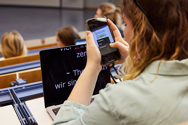 Eine Person mit lockigen Haaren hat auf ihren Handy eine Folie einer Präsentation abfotorafiert. Der Text ist markiert und kann durch ein Menüeintrag vorgelesen werden. Vor der Person steht zudem ein Laptop mit Notizen in größerer Schrift auf dem Bildschirm. Mehrer Personen sitzen um die Person herum.