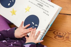 Von unten links kommende Kinderhände tasten eine Tastbilderbuchseite. Auf der Seite ist oben angeschnitten ein großgedruckter Text zu sehen. Darunter sind zwei gelbe Sterne und ein blauer Erdball. Der Erdball pustet drei weiße Wollfäden, die sich auf die nächste Buchseite schlängeln.