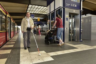Mann mit Langstock folgt Leitlinien und Noppenplatten am Bahnhof.