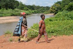 Ein Junge in einem Entwicklungsland führt eine ältere Person an einem Stock auf einem Weg vom Fluss.