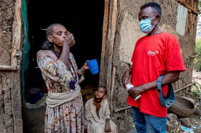 Eine afrikanische Frau steht vor dem dunklen Eingang einer ärmlichen Lehmbehausung und nimmt ein Medikament ein. Ein afrikanischer Mann mit rotem CBM-T-Shirt und Mundschutz, der die Medikamentenbox noch in der Hand hält und ein junges Mädchen im Hütteneingang sehen ihr dabei zu. 
