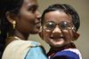 Ein kleines indisches Mädchen mit kurzen, schwarzen Haaren und einer Brille befindet sich auf dem Arm seiner Mutter. Das Mädchen blickt in die Kamera, der Blick der Mutter ist nach rechts gewandt.