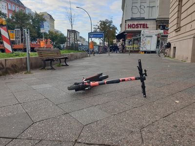 Ein E-Roller liegt mitten auf einem Gehweg in der Stadt. Links steht eine Bank. Im Hintergrund ist ein Hostel zu sehen.