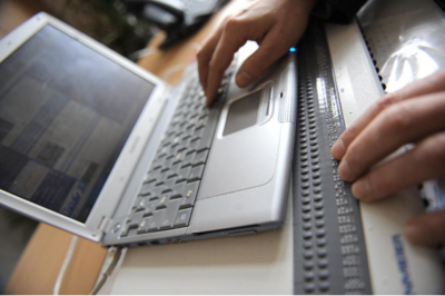 Auf der Tastatur eines Laptops liegt die rechte Hand einer Person. Die linke Hand liegt auf einer Braillezeile vor dem Laptop.