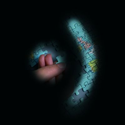 In ein Puzzle einer Weltkarte fügt eine Hand ein weiteres Puzzleteil ein. Der Großteil des Bildes ist schwarz, erkennbar ist nur ein kreisförmiger Ausschnitt der Hand und ein sichelförmiger Ausschnitt des Puzzles rechts davon.