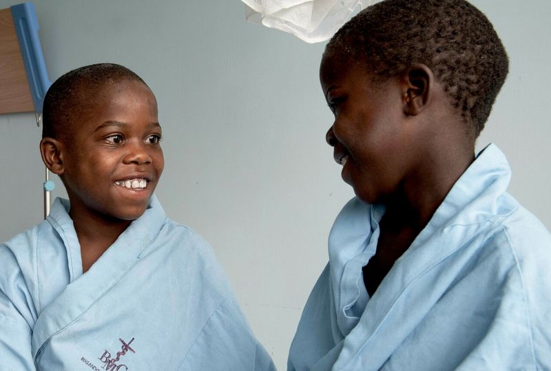 Nach der Operation im Krankenzimmer. Die acht- und zehnjährigen Brüder lächeln sich glücklich an. Sie sind in hellblaue viel zu große Krankenhauskittel gekleidet.