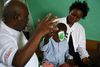 Foto eines kleinen afrikanischen Jungen bei der Augenuntersuchung nach der Operation am Grauen Star. Konzentriert ahmt er die Hand des Arztes nach (links von der Seite zu sehen), in dem er ebenso fünf Finger hebt. Sein anderes Auge ist noch zugeklebt. Hinter ihm sitzt seine Mutter und beobachtet zufrieden die Szene.