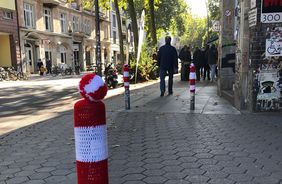 Auf einem belebten Bürgersteig steht im Vordergrund ein Poller mit rot-weiß-gestreifter Häkelmütze mit Bommel. Zwei weitere stehen dahinter. Dahinter laufen Menschen. Die Sonne scheint auf die Straße.