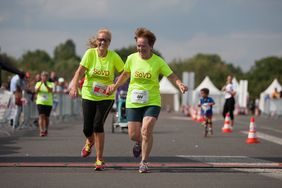 zwei Frauen in neon-grünen T-Shirts laufen Hand in Hand lachend über eine Ziellinie