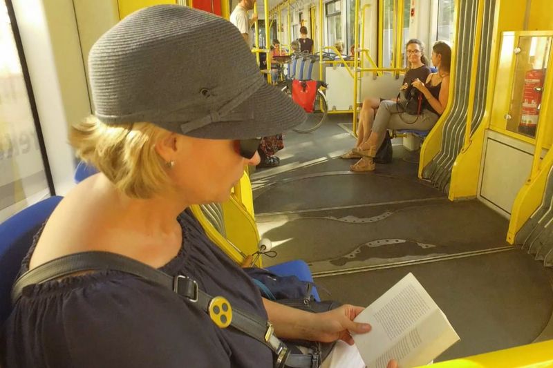 In der S-Bahn. Im Vordergrund sitzt eine sehbehinderte Frau und liest in einem Buch. Im Hintergrund reden zwei Frauen mit Blick auf die sehbehinderte Frau.