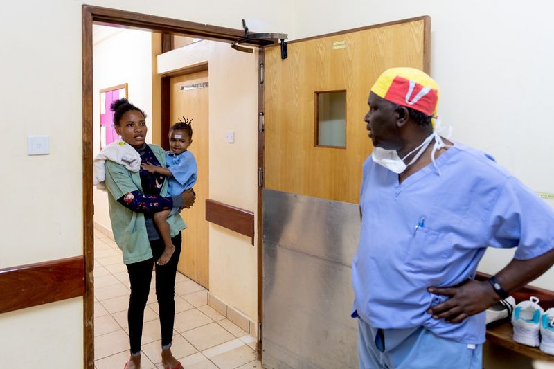 Eine junge Mutter steht mit ihrem Kleinkind im Türrahmen eines Krankenhauses. Ihr gegenüber steht eine Pflegekraft.
