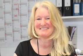 Portraitbild einer Frau mit blonden langen Haaren. Sie trägt ein schwarzes T-Shirt und lächelt in die Kamera. Im Hintergrund sind Büroutensilien zu sehen.