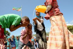 Ein Person gießt Wasser aus einem Kanister. Kinder fangen das Wasser mit den Händen auf und waschen sich das Gesicht.