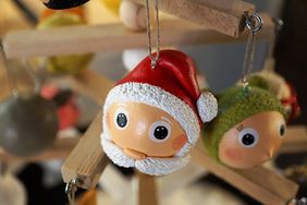An einem stilisierten Weihnachtsbaum aus Holzlatten hängt eine Weihnachtsmann-Holzfigur. Diese besteht nur aus einer Kugel als Kopf mit roter Mütze und Bart. Sie hat große runde Augen. Im Hintergrund rechts hängt eine ähnliche Figur mit grüner Mütze. Links hinten hängt eine graue Kugel.