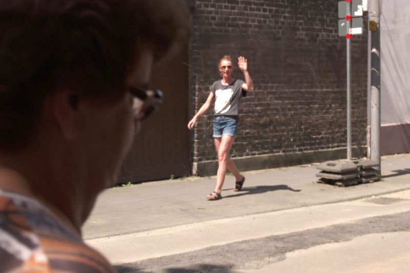 Auf der Straße grüßt eine Frau auf der anderen Straßenseite eine im Vordergrund des Bildes gehende Frau.