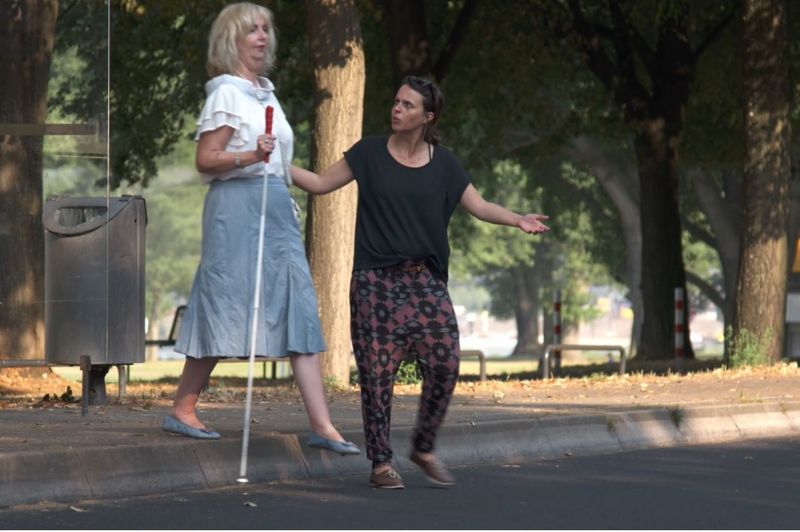 Eine Frau zieht am Arm einer sehbehinderten Frau mit Stock und weist über die Straße.