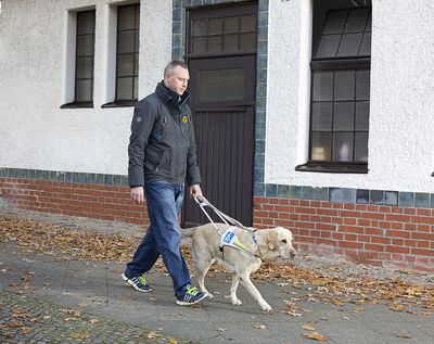 Ein Mann läuft mit seinem Blindenführhund auf dem Gehsteig vor einem Haus.