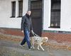 Ein Mann läuft mit seinem Blindenführhund auf dem Gehsteig vor einem weißen Hausfassade.