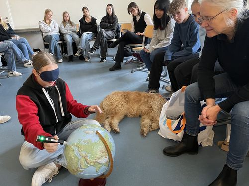 Ein Mädchen im Alter von 14 Jahren sitzt in einem Klassenraum auf dem Boden und ertastet einen taktilen Globus. Neben ihr ein Blindenführhund und die Besitzerin dessen. 
