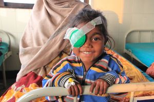 Ein kleines lachendes Mädchen mit bunt getreifter Jacke schaut über das Fußende eines Klinikbetts. Das rechte Auge des Mädchen ist mit einem Verband zugeklebt. 
