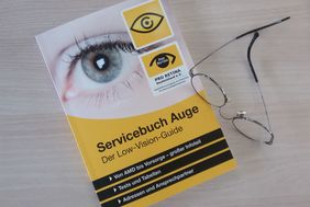 Oben auf dem diagonal liegenden Buch ist ein großes Auge abgebildet, darunter schwarz auf gelb der Titel "Servicebuch Auge - Der Low-Vision-Guide". Rechts auf dem Buch liegt eine silberne Drahtgestellbrille.