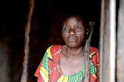 Flussblindheit – erblindete Frau Eine ältere erblindete Frau steht mit einem Stock vor einem dunklen Hintergrund.