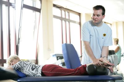 Ein blinder Physiotherapeut behandelt eine Frau an der Ferse. Die Frau liegt auf einer Liege, der Physiotherapeut steht daneben.