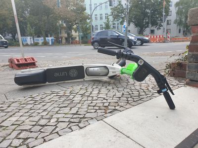 Ein E-Roller liegt auf dem Gehweg an einer befahrenen Straßenkreuzung.