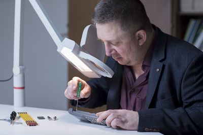 Ein Mann mittleren Alters mit kurzen, dunkelgrauen Haaren arbeitet mit einem Schraubenzieher an einem flachen elektronischen Gerät. Dabei schaut er durch eine beleuchtete Lupe, die an einem schwenkbaren Arm vor seinem Gesicht angebracht ist.
