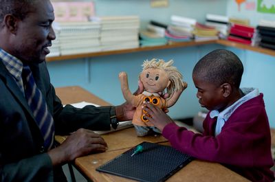 Ein Lehrer sitzt mit einem Junge zusammen, der an einer Puppe mit Punkten der Brailleschrift tastet.