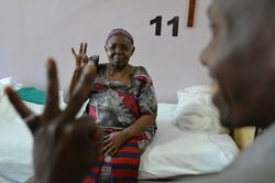 Grauer Star – Sehtest Eine ältere Frau sitzt auf einem Bett. Sie macht einen Sehtest und ahmt die Anzahl der Finger nach, die sie gezeigt bekommt.