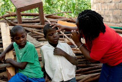 Eine afrikanische Frau mit rotem T-Shirt und blauem Rock steht rechts neben zwei kleinen Jungen in abgewetzter Kleidung, die sich schüchtern an einen Holzstapel lehnen. Dem näherstehenden Jungen hat sie das Augenlid leicht angehoben und leuchtet ihm mit einer Taschenlampe ins Auge. 
