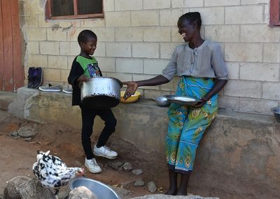 Ein afrikanischer Junge schleppt zufrieden schmunzelnd einen großen metallenen Kochtopf zu seiner Mutter, die ihn begeistert anlacht und die Hand ausstreckt, um ihm den Topf abzunehmen.  
