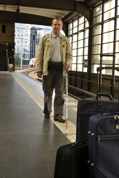Mann mit Langstock folgt Leitlinien am Bahnhof. Gepäckstücke blockieren den Weg.
