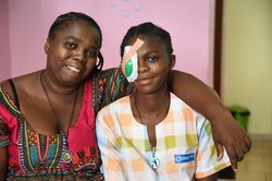 Ein Mädchen nach einer Augenoperation mit verbundenem Auge sitzt neben ihrer Mutter. Beide lächeln.
