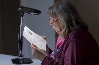 Eine Frau mittleren Alters mit schulterlangen, grauen Haaren liest ein bedrucktes Blatt Papier, das sie unter eine Leseleuchte hält. Sie trägt Oberbekleidung in Dunkelviolett und ein dazu passendes Halstuch.