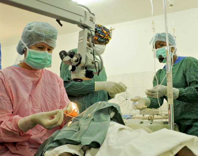 Dr. Irmela Erdmann bei einer Augenoperation. Sie trägt Haar-, Mundschutz und OP-Handschuhe. Der Patient ist abgedeckt bis auf das zu operierende Auge. Zwei afrikanische OP-Pflegerinnen, ein Mann und eine Frau bereiten Infusionen vor. 