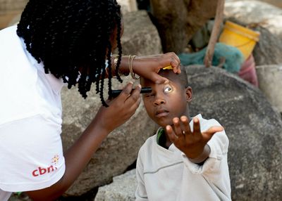 Eine afrikanische Frau mit geflochtenen Zöpfen beugt sich mit einer Taschenlampe über einen afrikanischen Jungen, hebt sein Augenlid an und leuchtet ihm in ein Auge. 