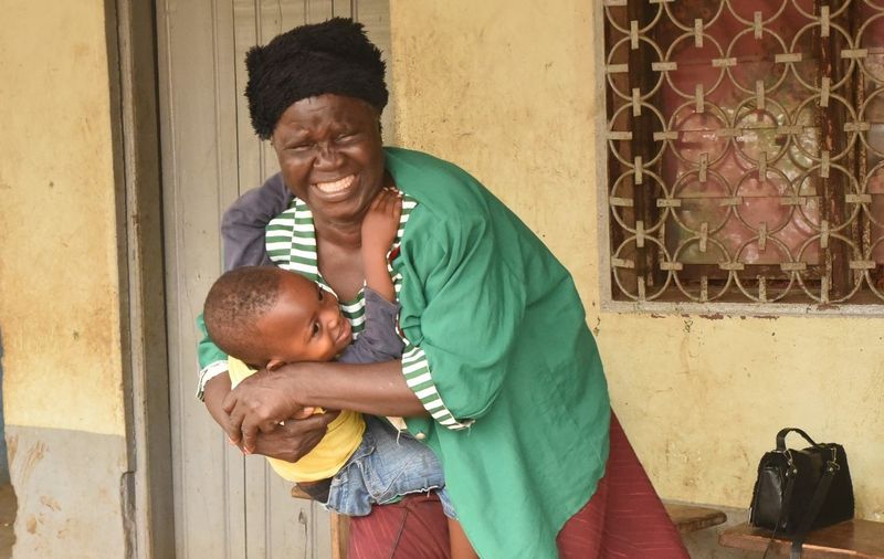 Eine afrikanische Frau mit grüner Jacke und rotem Rock umarmt vor Glück beinahe weinend ihren kleinen Enkel, der sich vor Freude strahlend an sie klammert. 