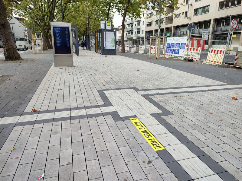 Gepflasterte Straße mit Leitstreifen für blinde Menschen und einem großen gelben Aufkleber "Bitte Weg frei!"