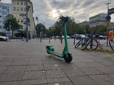 Ein E-Roller steht mitten auf dem Gehweg an einer befahrenen Kreuzung in der Stadt. Autos und Radfahrer fahren im Hintergrund über die Kreuzung.