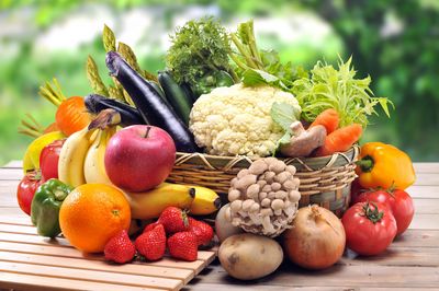 Ein Korb voller Gemüse auf einer Holzplatte. Um den Korb verteilt verschiedenes Obst.