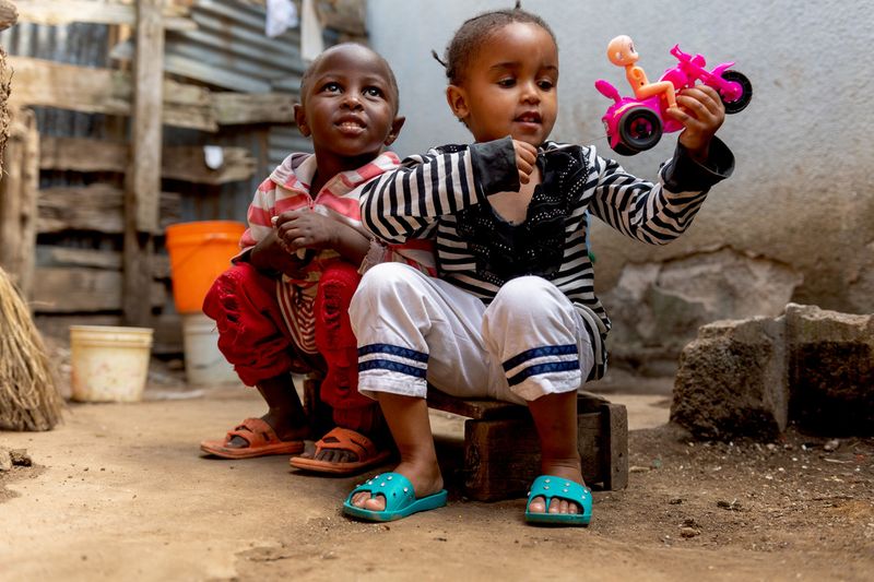 Zwei Kleinkinder sitzen auf einer kleine Holzbank, eines der Kinder hält ein Spielzeug in der Hand.