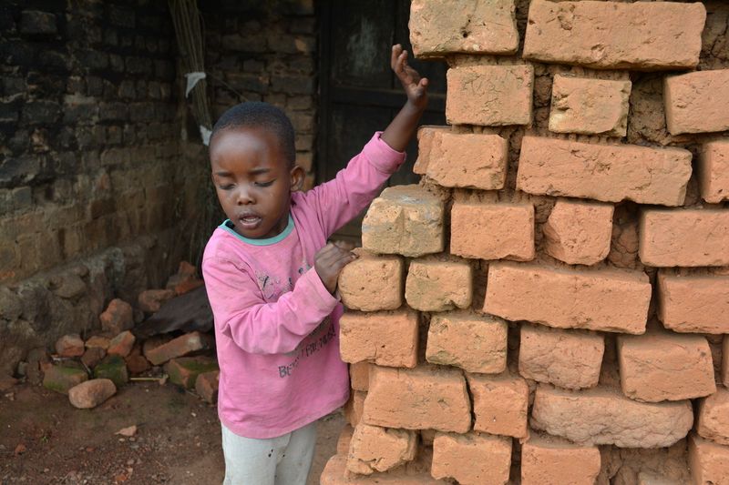 Ein kleiner Junge aus einem Entwicklungsland steht am Rand einer Ziegelmauer. Er tastet nach den Steinen.