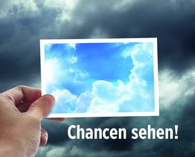 Motiv 2016: Es zeigt einen grauen, wolkigen Himmel. Im Vordergrund hält eine Hand vom linken unteren Bildrand her eine Postkarte in die Bildmitte. Die Karte zeigt einen blauen Himmel mit weißen Wolken. Unter der Postkarte steht das Motto „Chancen sehe